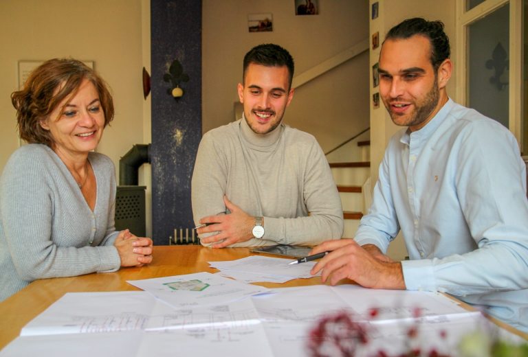 Zwei Männer und eine Frau sitzen gut gelaunt an einem Tisch mit Gebäudeplänen.